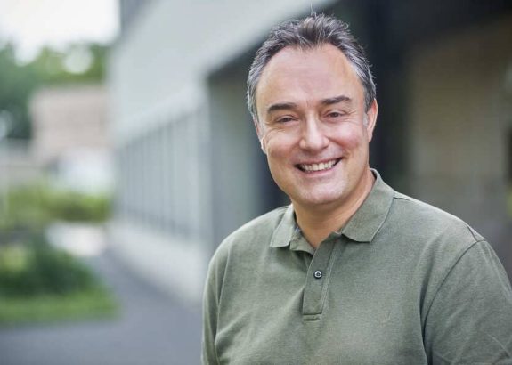 Portret van een lachende man met kort grijzend haar in een olijfgroen poloshirt, buiten voor een modern gebouw met zachte, groene achtergrond.
