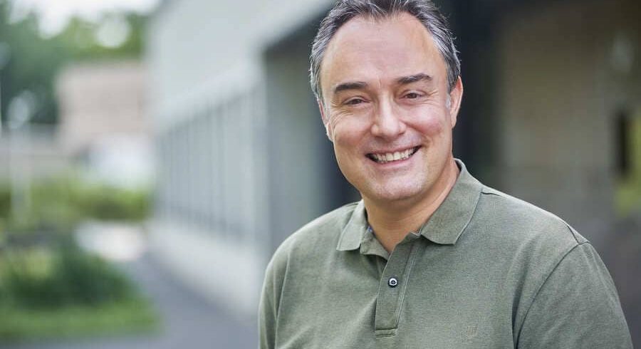Portret van een lachende man met kort grijzend haar in een olijfgroen poloshirt, buiten voor een modern gebouw met zachte, groene achtergrond.